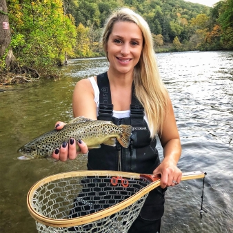 A woman standing in river holding a fish over a net (Instagram@theblondeangler)